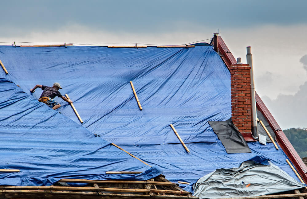 The roofer works on roof when is rain. The tarp covers the roof of the old house in the reconstruction.